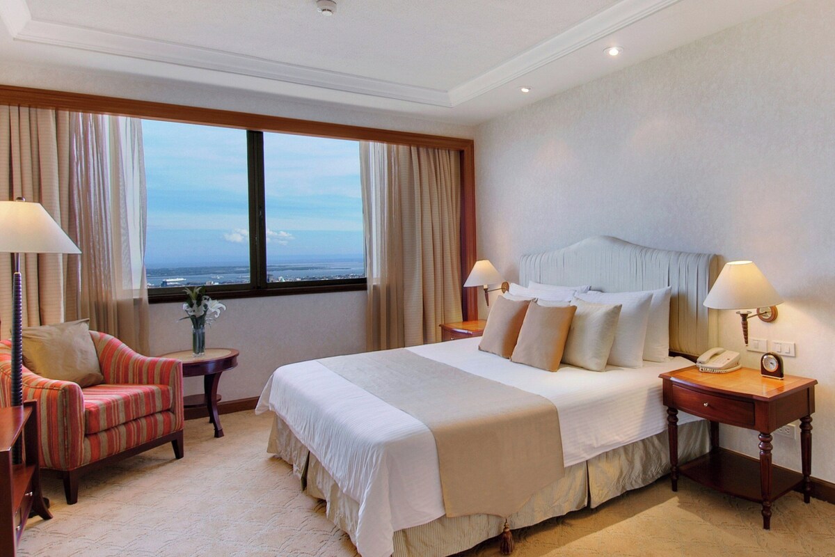 Een hotelkamer met een groot bed, twee nachtkastjes, een stoel, een bureau en uitzicht op de oceaan.