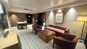 Camera d'hotel con tappeto a righe, un divano, una poltrona, un tavolino e una televisione.