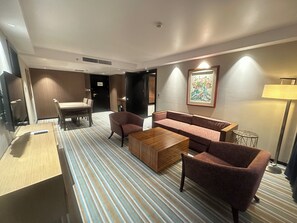 Habitación de hotel con alfombra a rayas, un sofá, una silla, una mesita y un televisor.