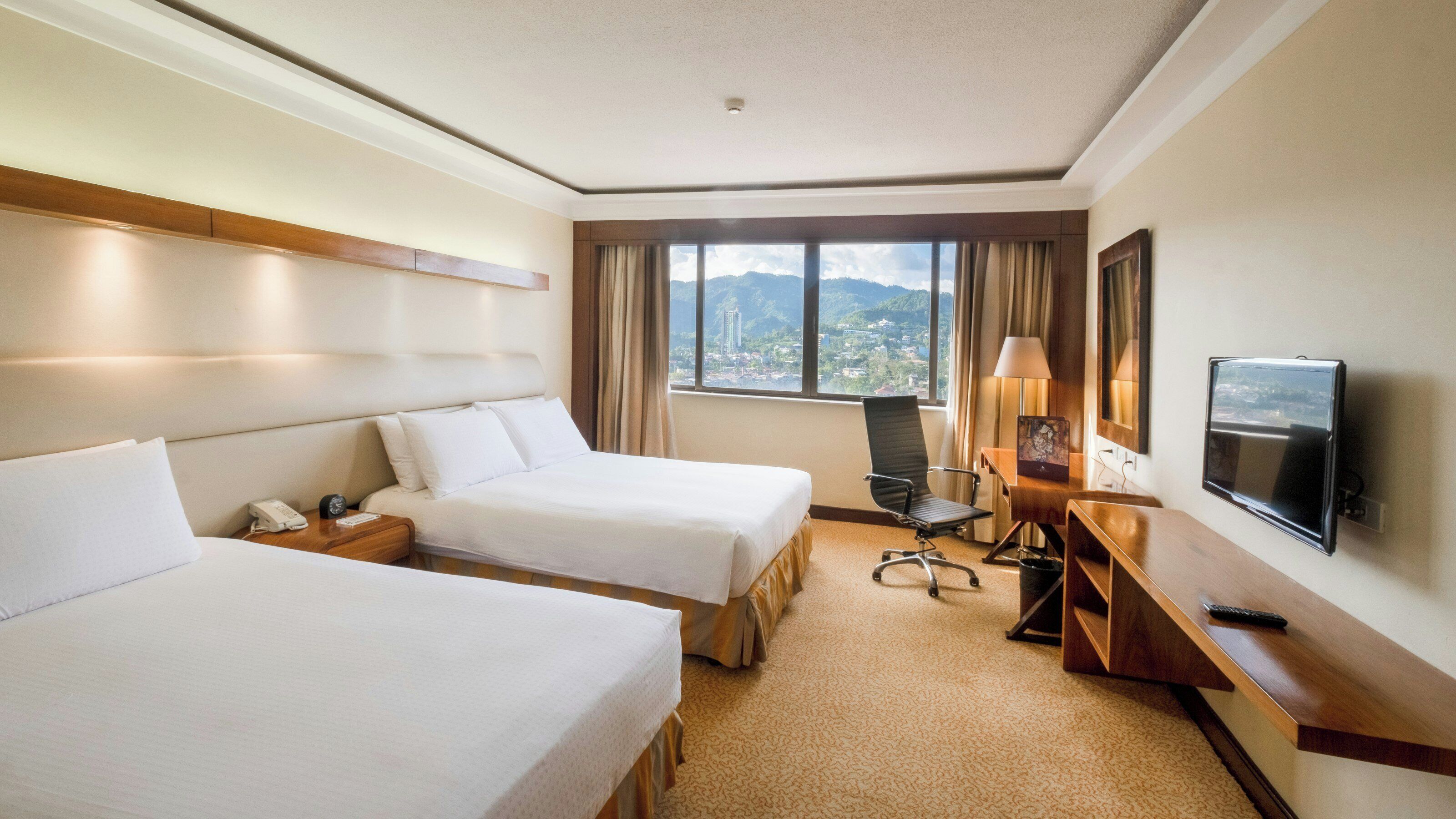 A hotel room with two beds, a desk, a chair, a TV, and a window with a mountain view.