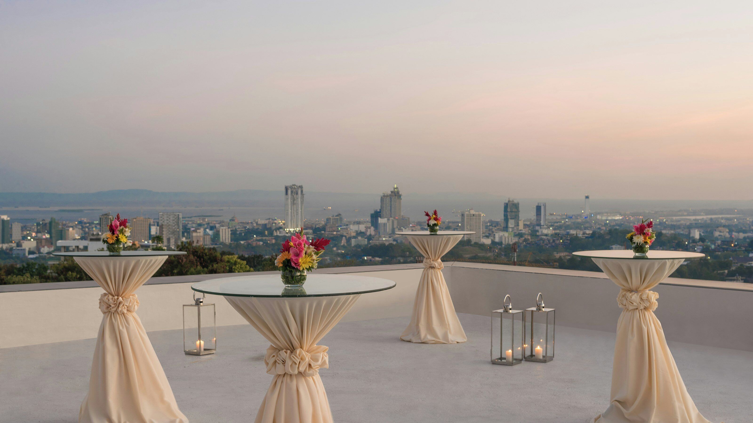 A rooftop terrace with four round tables, each with a vase of flowers and candle lanterns, overlooking a cityscape.