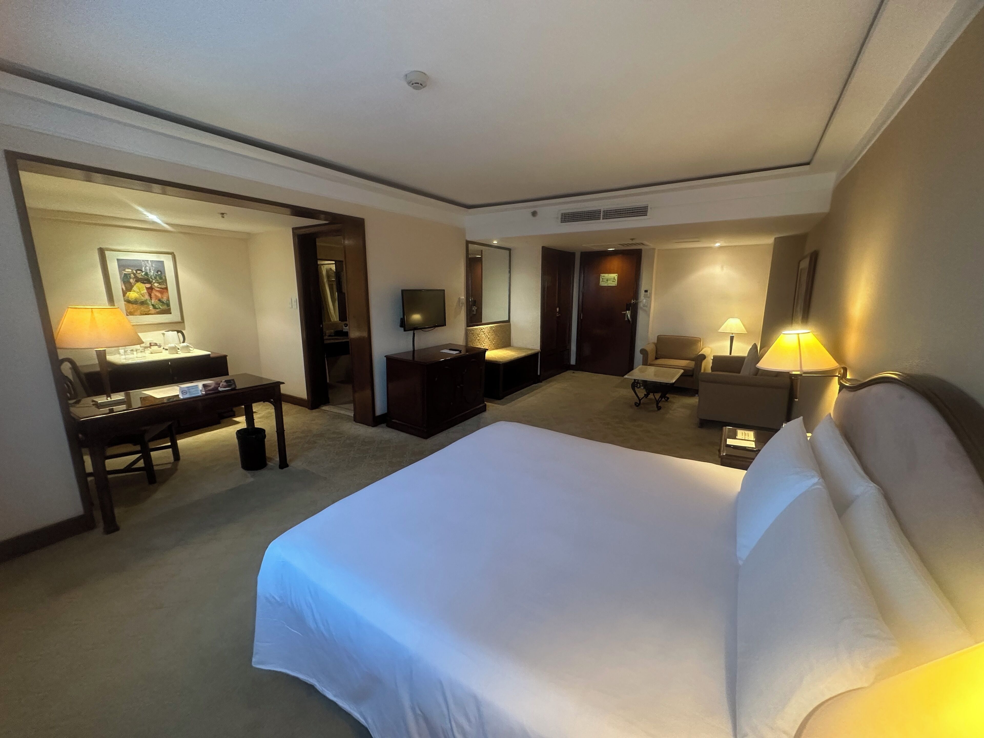 A hotel room with a large bed, a desk with a lamp, a TV, and a chair.
