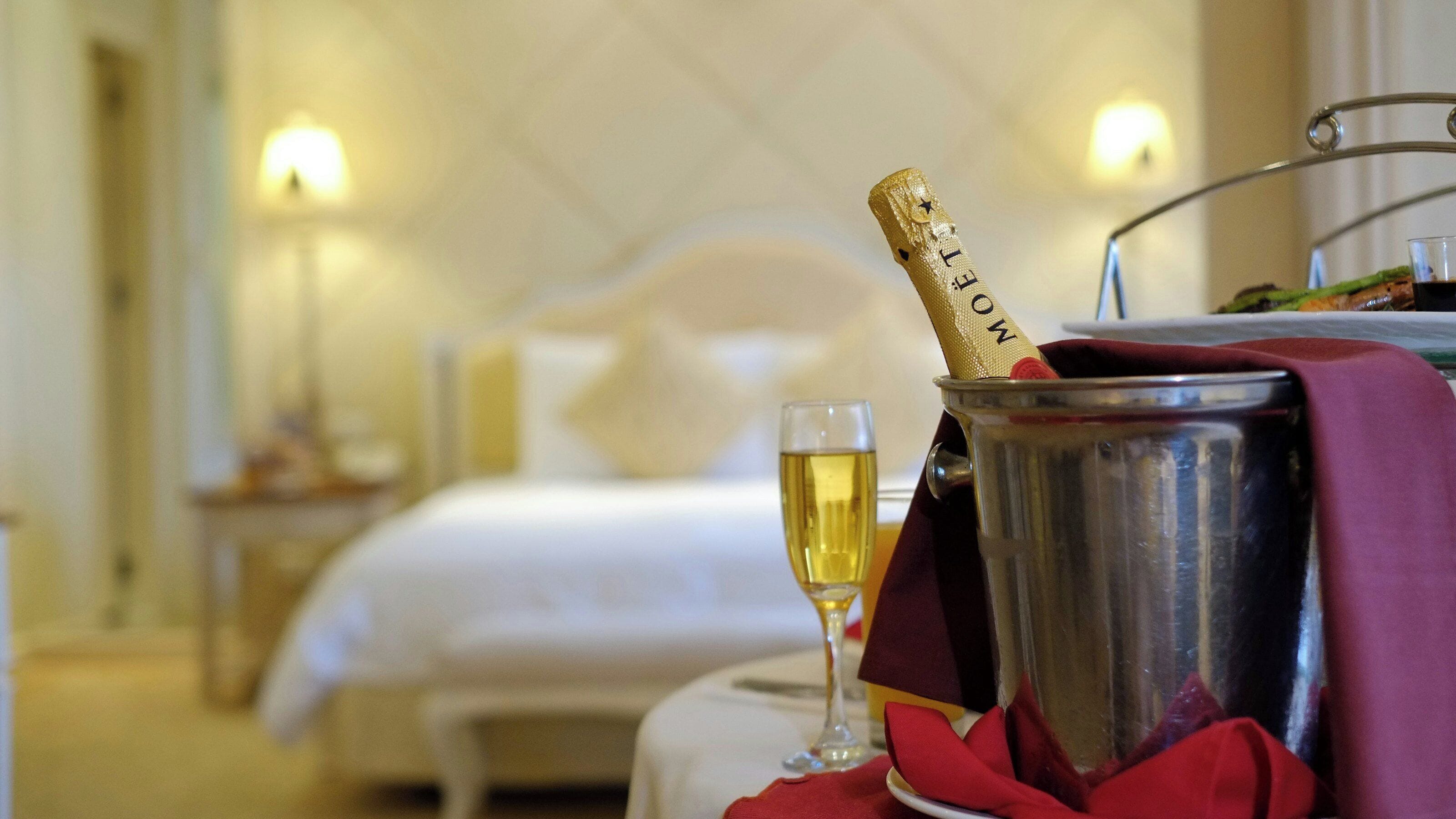 A bed with a white headboard, a bedside table with a glass of champagne, and a metal bucket with a corkscrew.