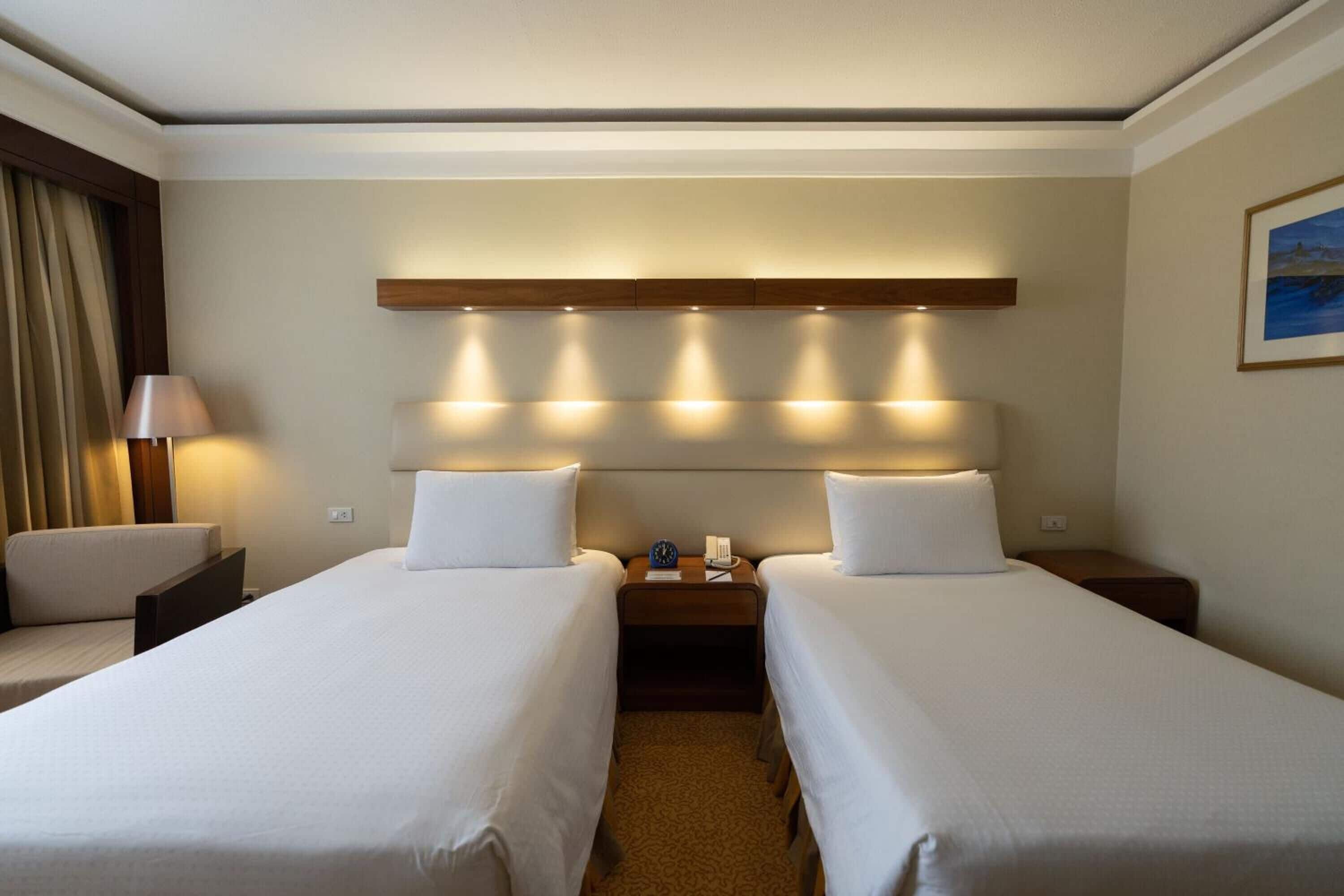 Habitación de hotel con dos camas, una mesita de noche con lámpara y cabecero con iluminación integrada.