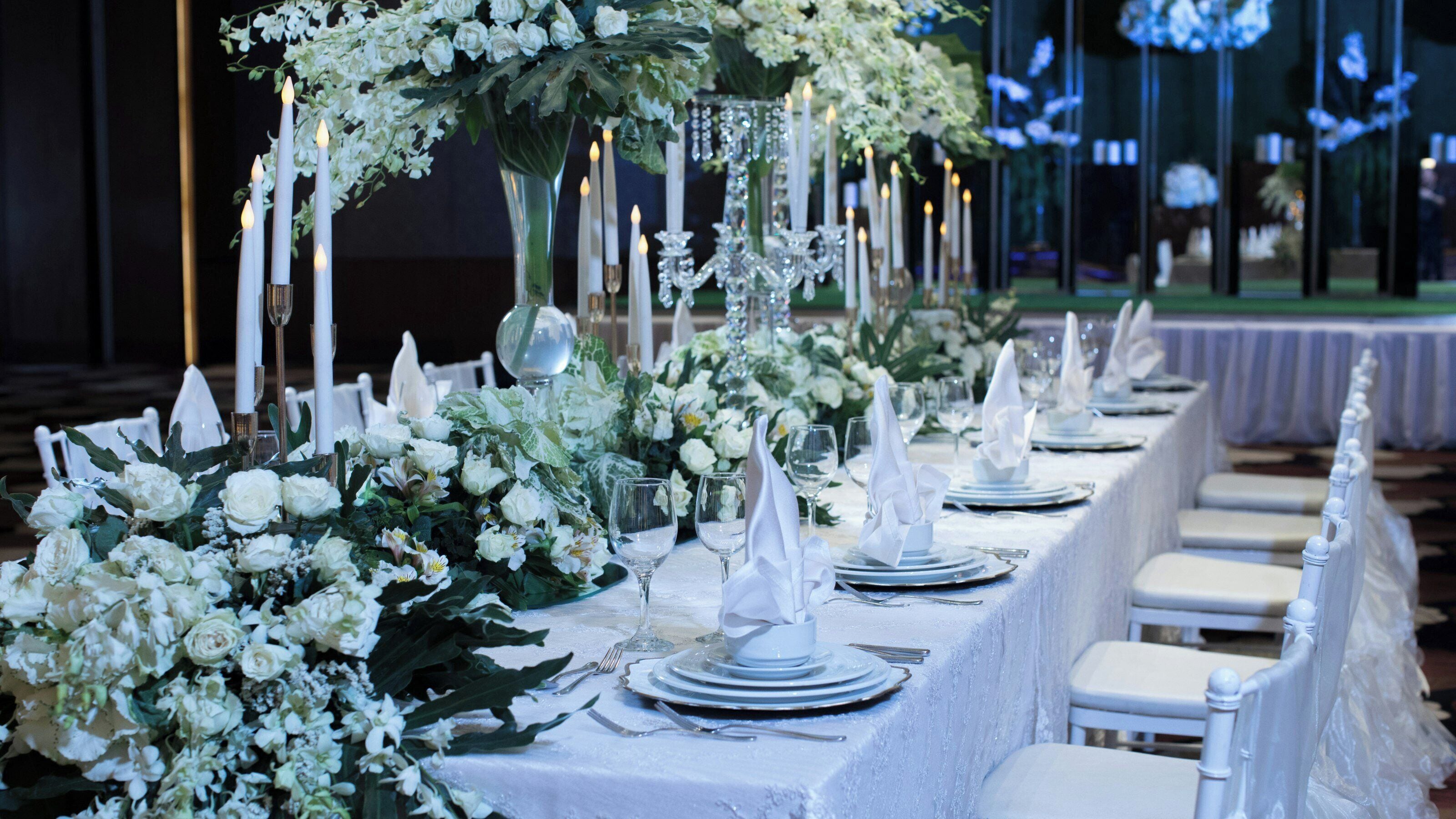 A long banquet table set for a formal event with white tablecloths, glassware, and candle holders.
