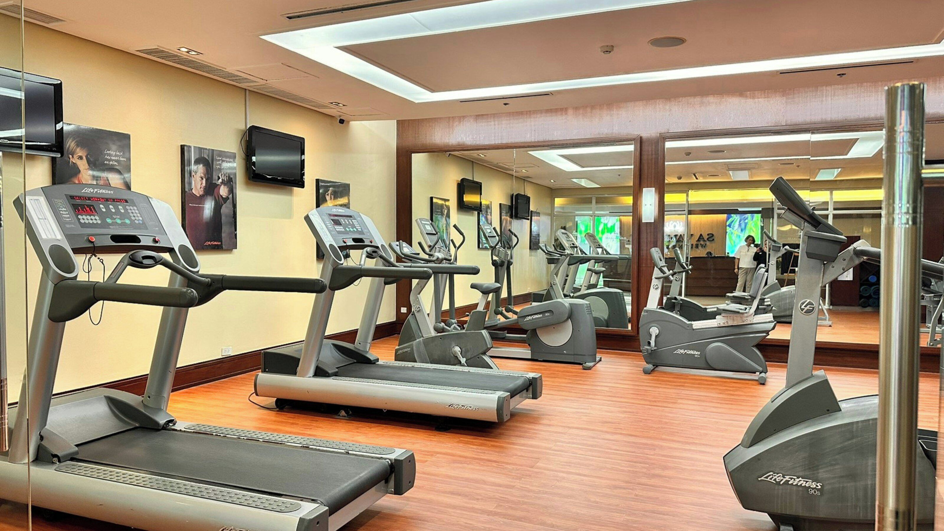 A gym with multiple treadmills and elliptical machines, a wooden floor, and a mirror reflecting the equipment.