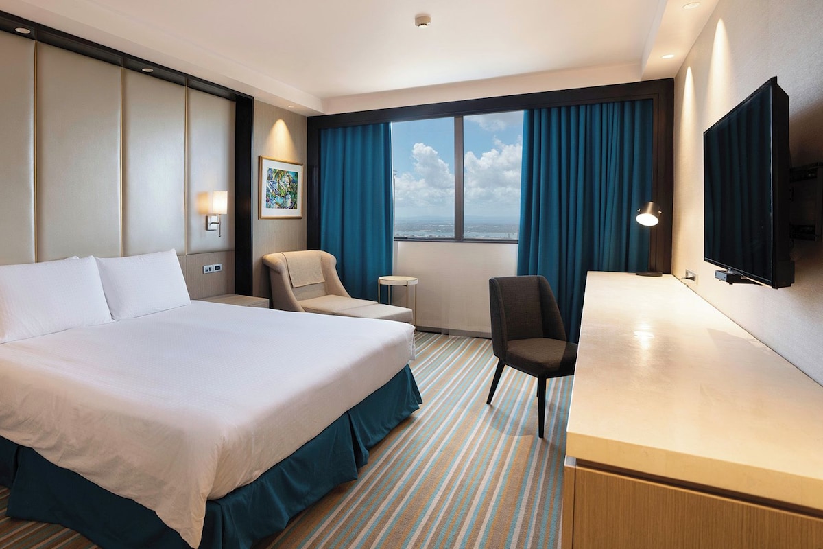 A hotel room with a large bed, a desk, a chair, a TV, and a window with curtains.