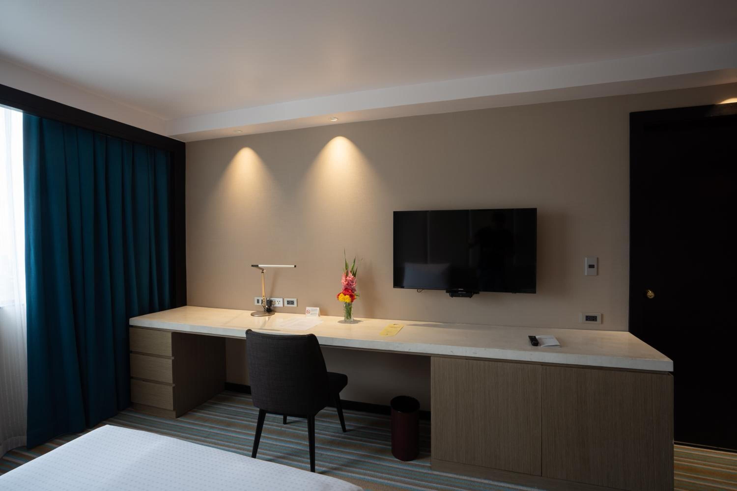 A hotel room with a bed, a desk with a chair, a TV mounted on the wall, and a window with curtains.