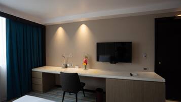 A hotel room with a bed, a desk with a chair, a TV mounted on the wall, and a window with curtains.