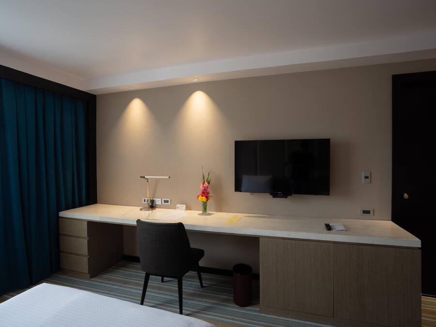 A hotel room with a bed, a desk with a chair, a TV mounted on the wall, and a window with curtains.