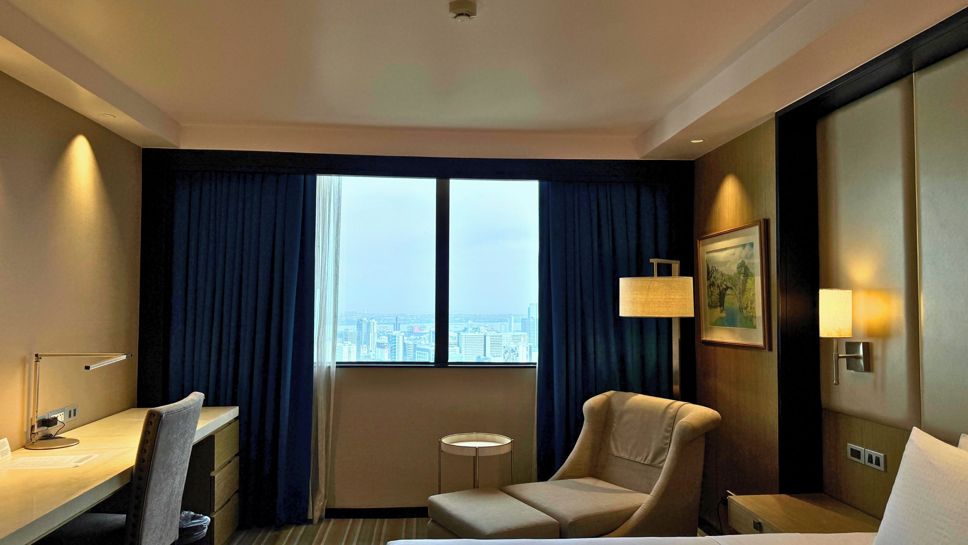 A hotel room with a desk, chair, bed, bedside table, lamp, and a city view through the window.