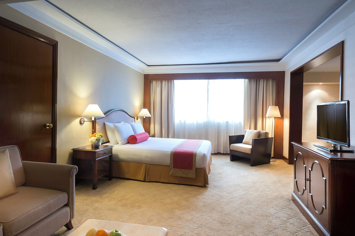 Una habitación de hotel con cama, televisión, escritorio, silla y ventana con cortinas.