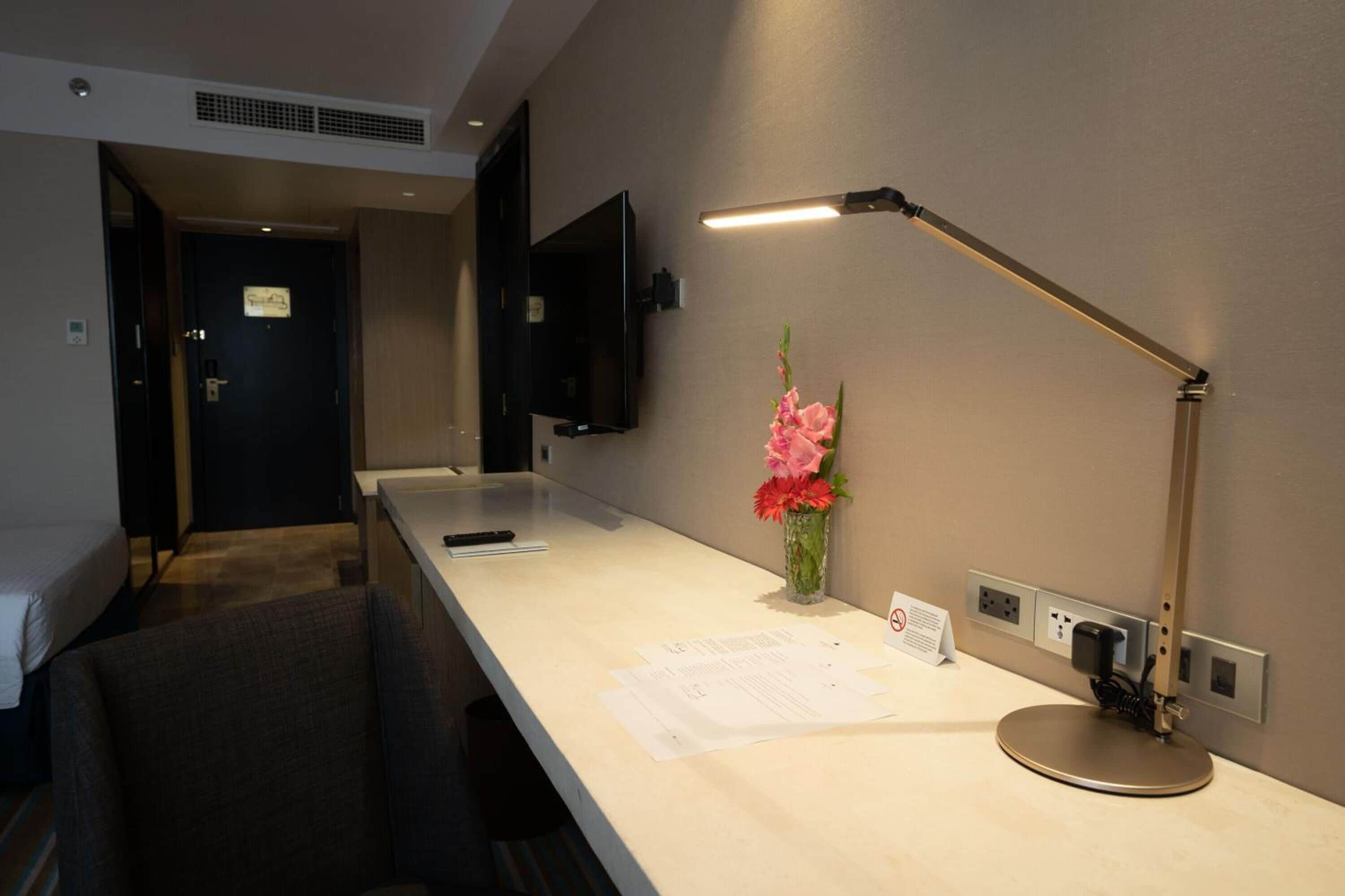 A hotel room with a bed, a desk with papers, a desk lamp, a flower vase, and a TV mounted on the wall.