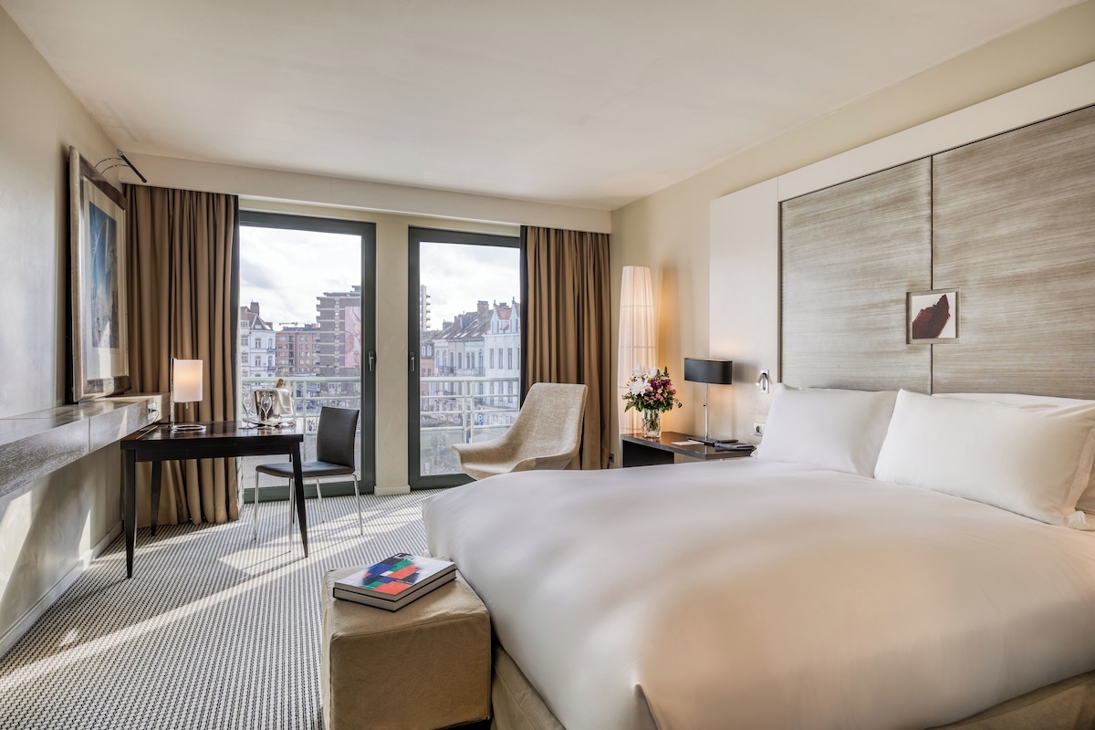 A hotel room with a large bed, a desk, a chair, a nightstand, and a view of the city.