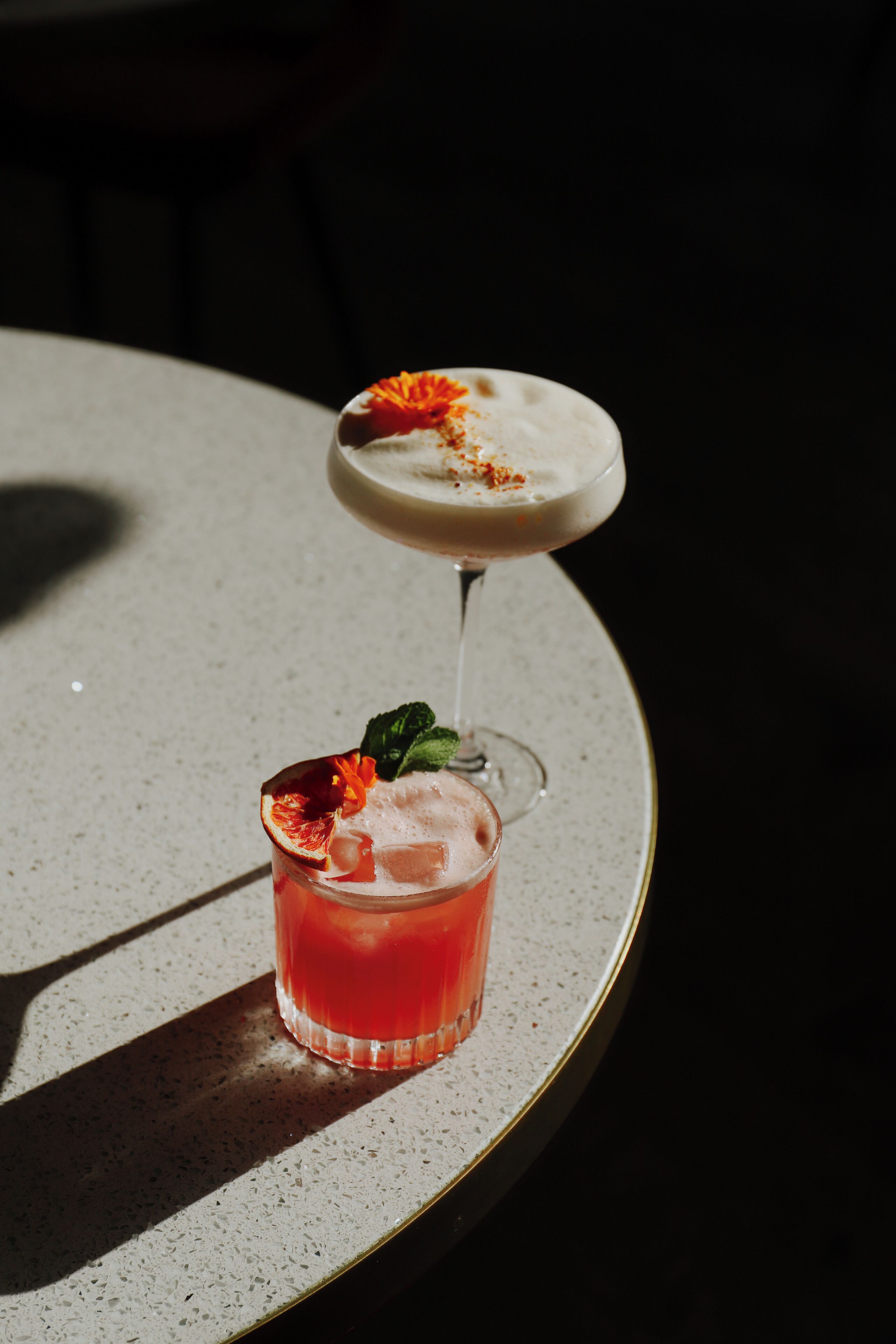 A round table with two glasses of cocktails, one with a garnish and one with a frothy top.
