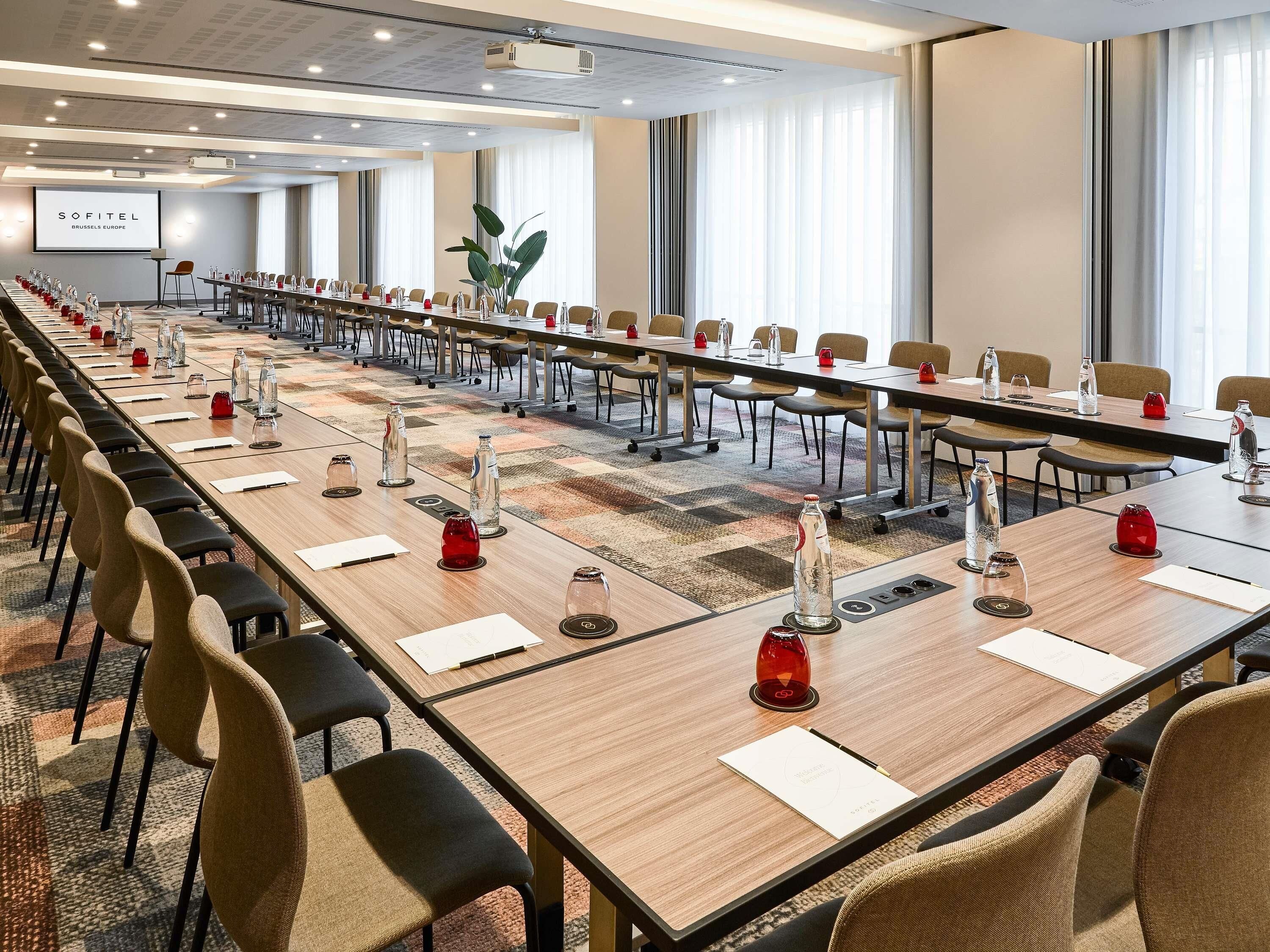 Un salón de conferencias con mesas largas, sillas, botellas de agua y una pantalla de proyector.