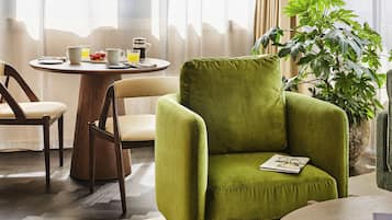 A green armchair with a magazine, a wooden table with a coffee maker and cups, and a large plant in a room with a herringbone floor.