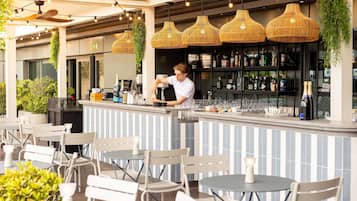Un área de bar al aire libre con un barman, mesas redondas, sillas, lámparas de mimbre colgantes y una barra con estantes llenos de botellas.