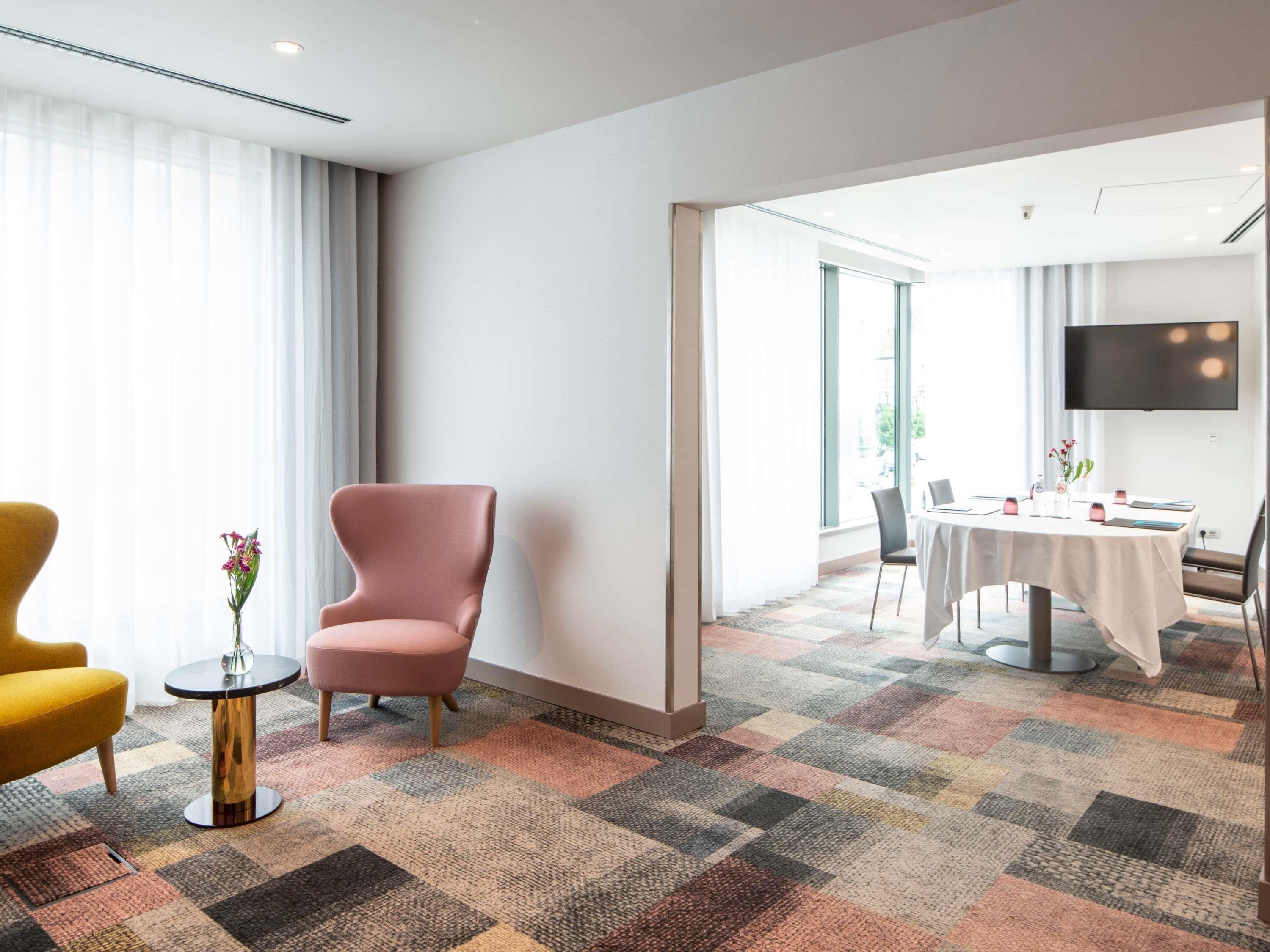 Una habitación de hotel con un ventanal grande, un sillón rosa, una mesita redonda y un comedor con una mesa y sillas.