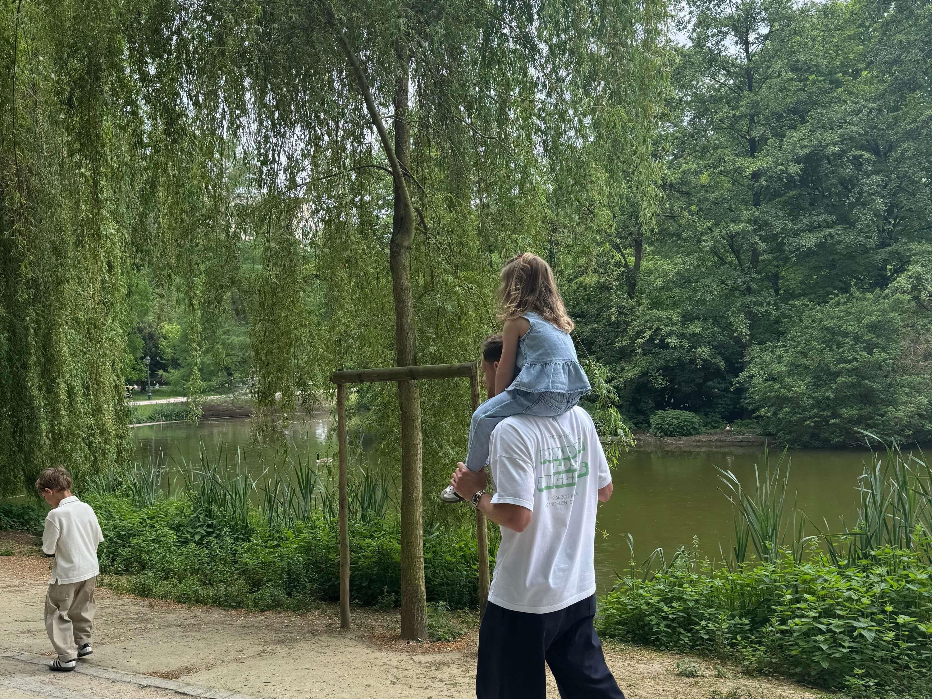 A man carrying a child on his shoulders near a body of water with trees and greenery in the background.