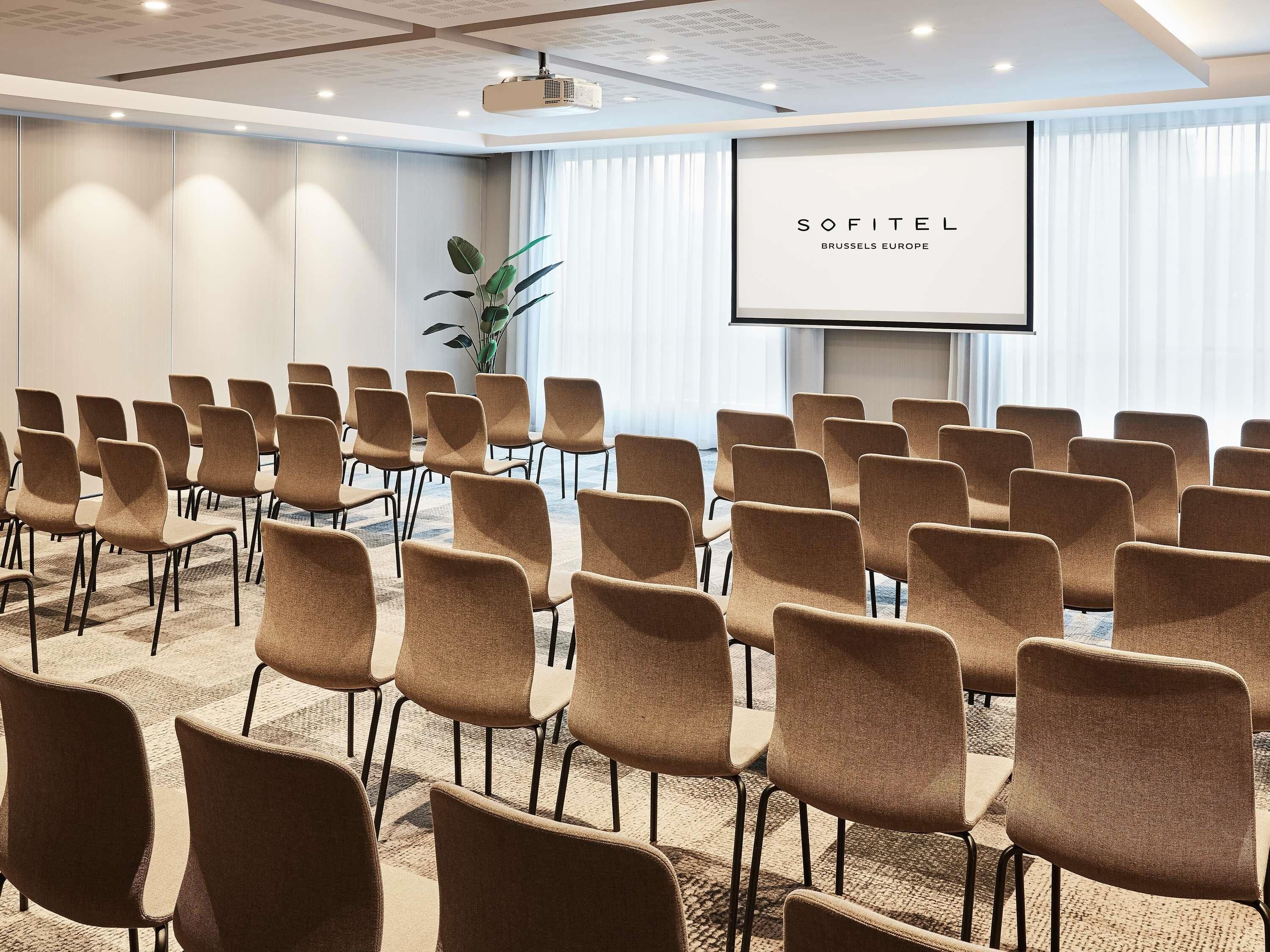 Una sala de conferencias con filas de sillas, una pantalla para proyector y una planta.