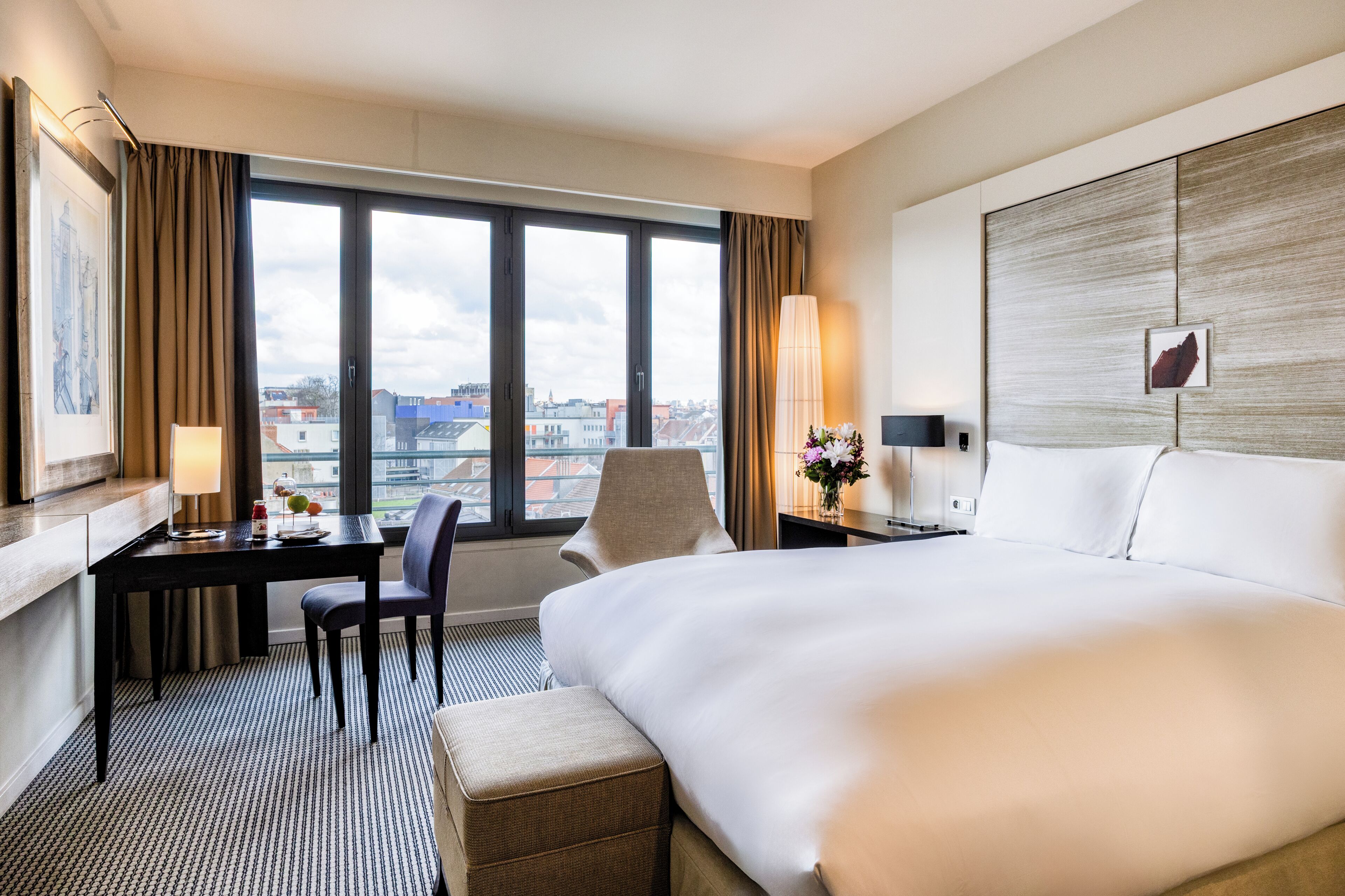 Habitación de hotel con una cama grande, un escritorio con una silla, una lámpara y vistas de la ciudad a través de la ventana.