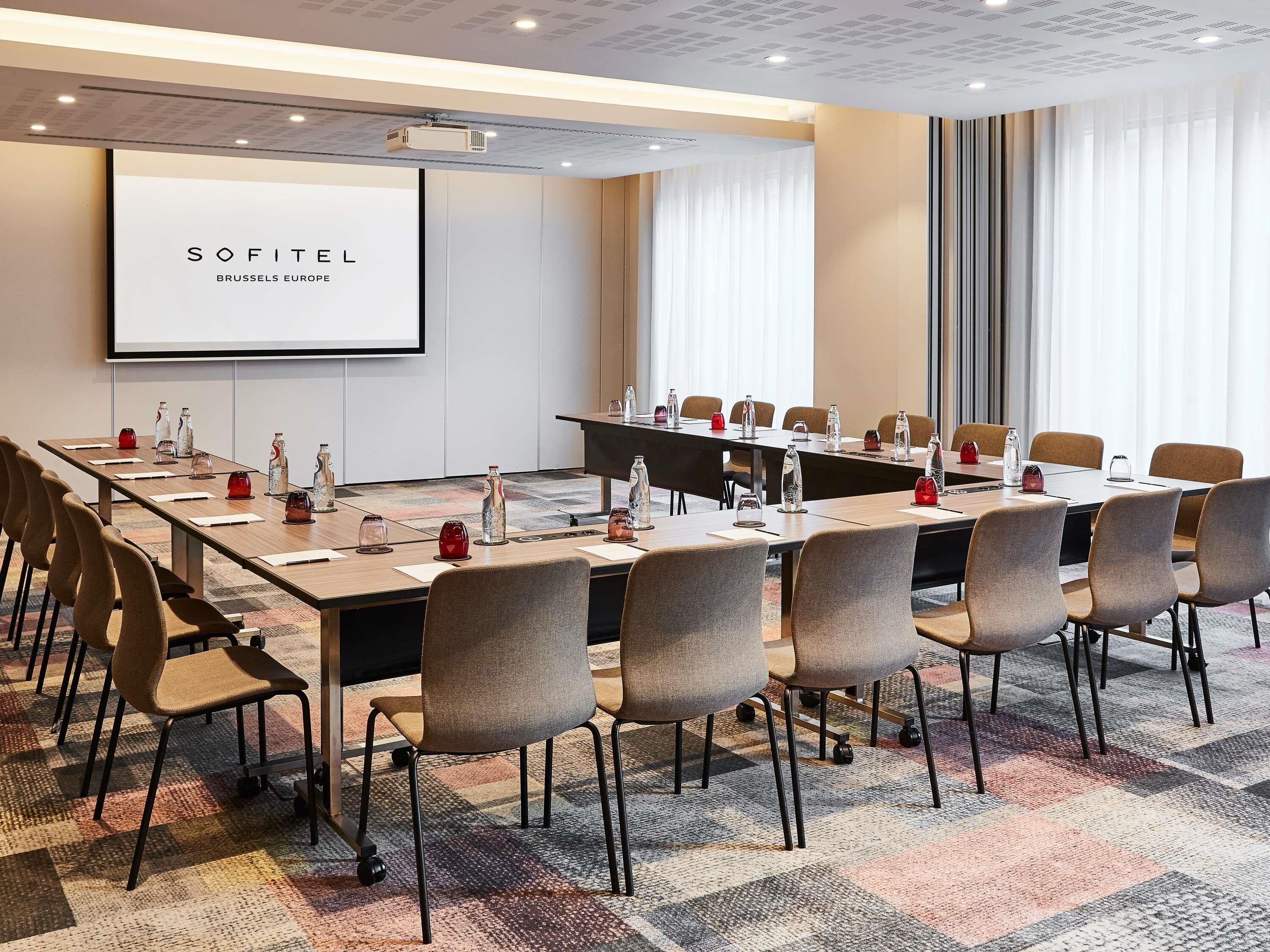 A conference room with a projector screen, long tables, and chairs, featuring a 'SOFITEL BRUSSELS EUROPE' sign.