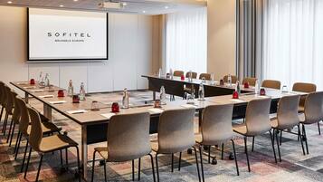 A conference room with a projector screen, long tables, and chairs, featuring a 'SOFITEL BRUSSELS EUROPE' sign.