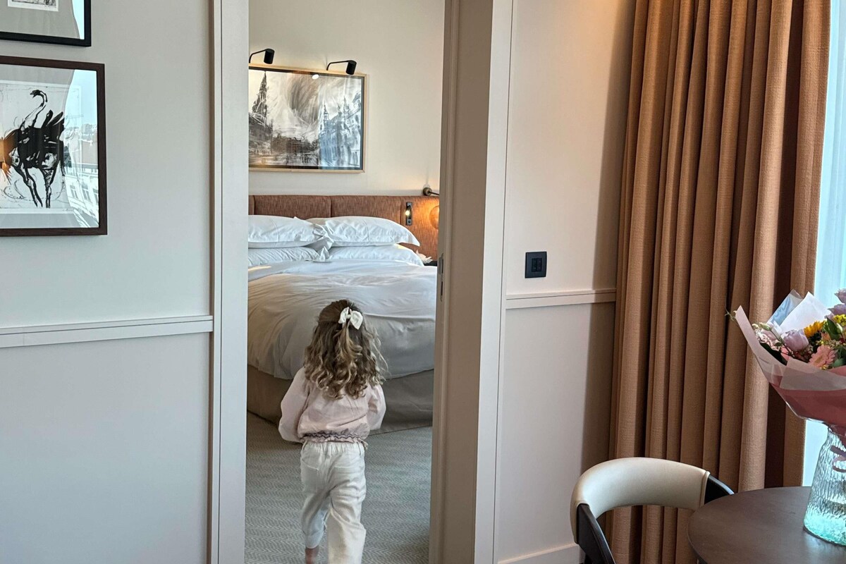 A hotel room with a bed, a painting on the wall, a curtain, and a table with a vase of flowers.