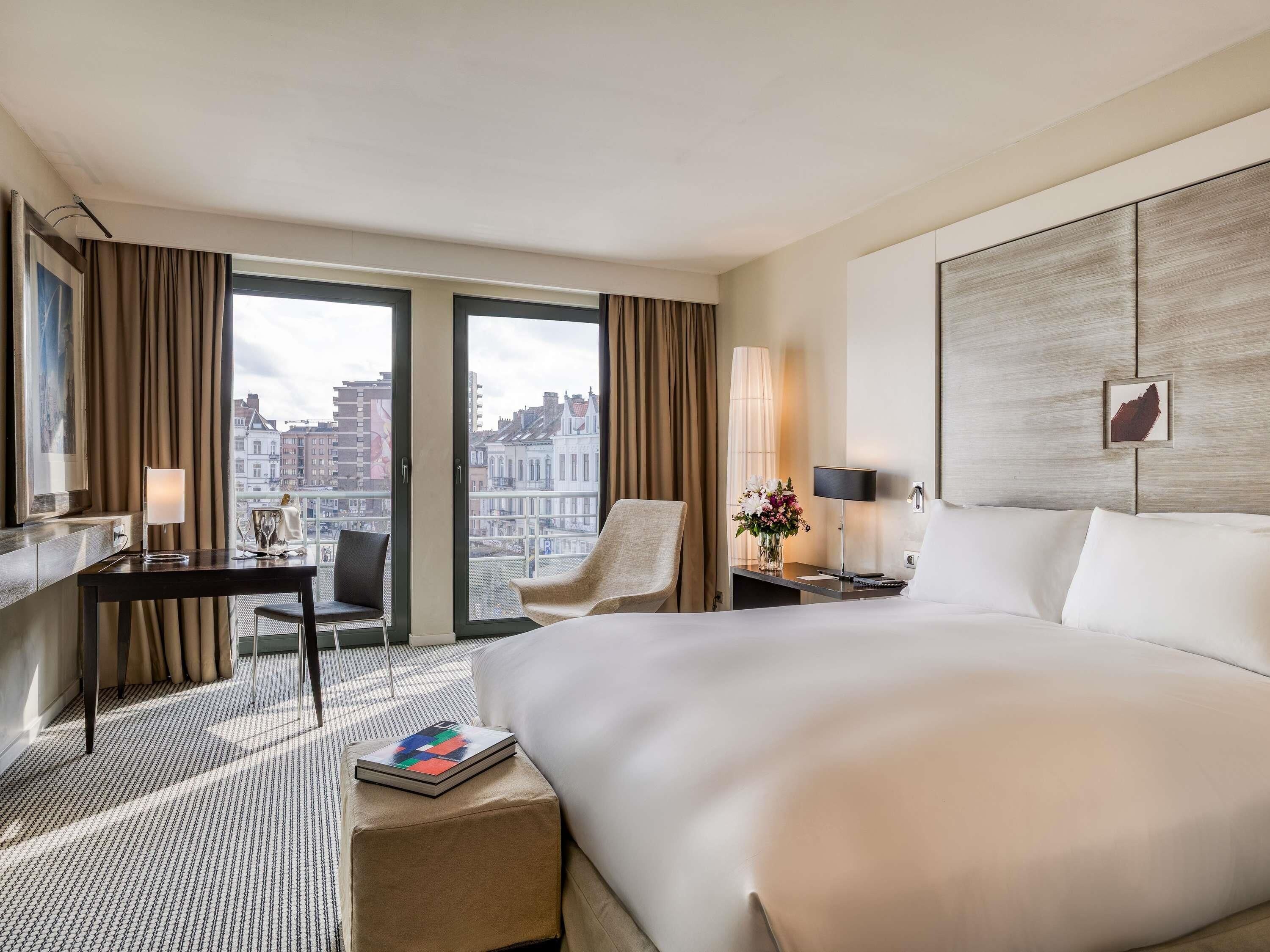 A hotel room with a large bed, a desk, a chair, a small table, and a view of the city.