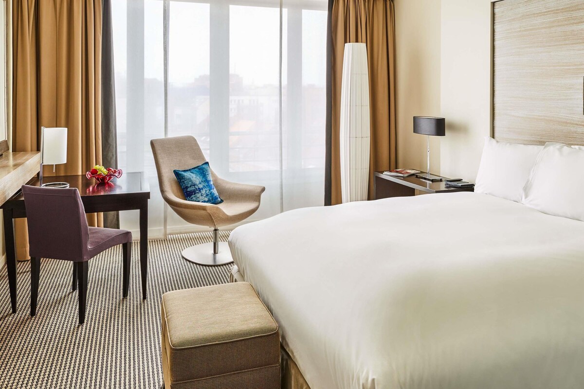 A hotel room with a bed, a desk with a chair, a lamp, and a view of the city.