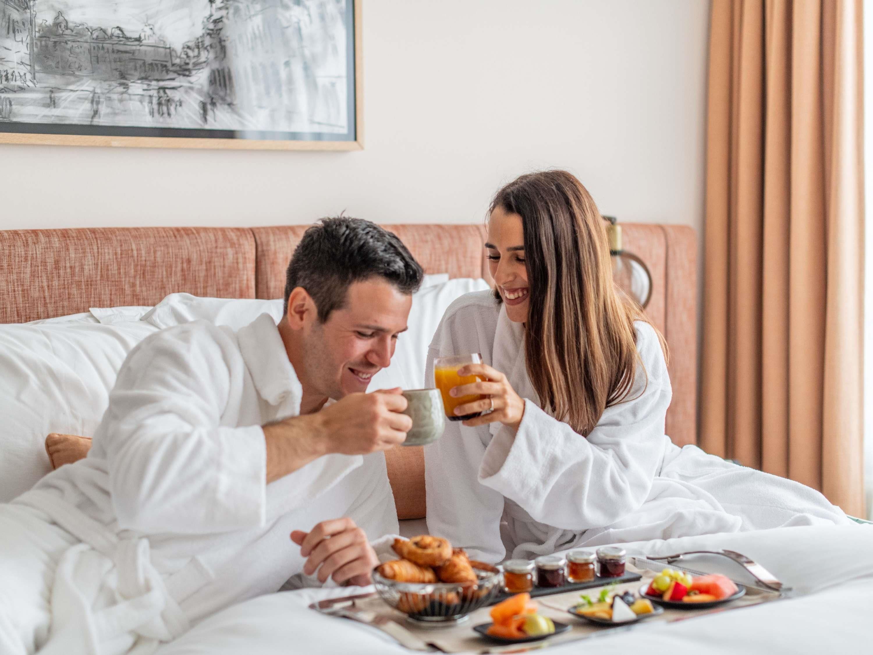 Una pareja en una habitación de hotel, sentada en la cama con comida para el desayuno y un cuadro en la pared.