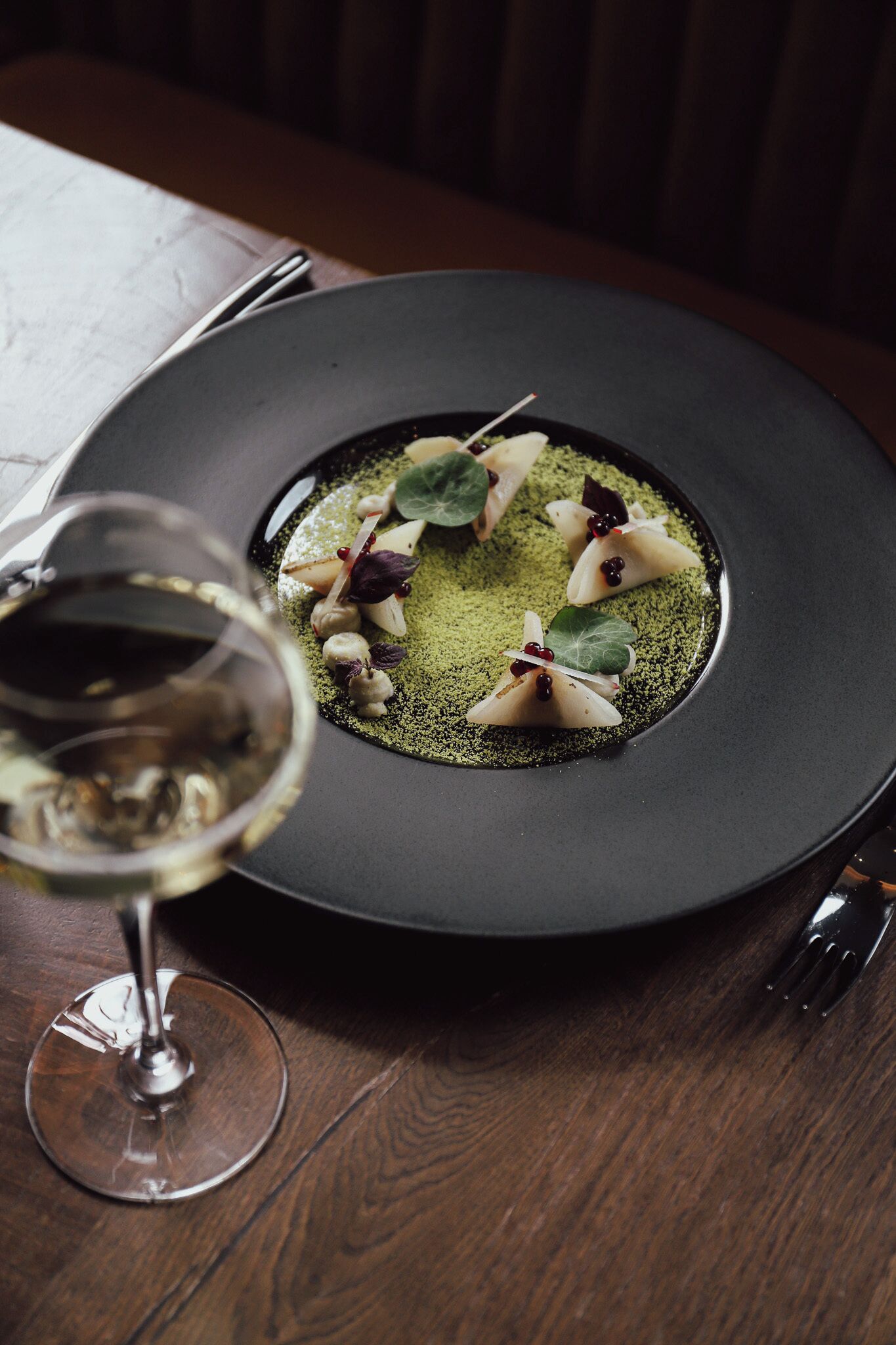 A black plate with a green garnish and a glass of white wine on a wooden table.