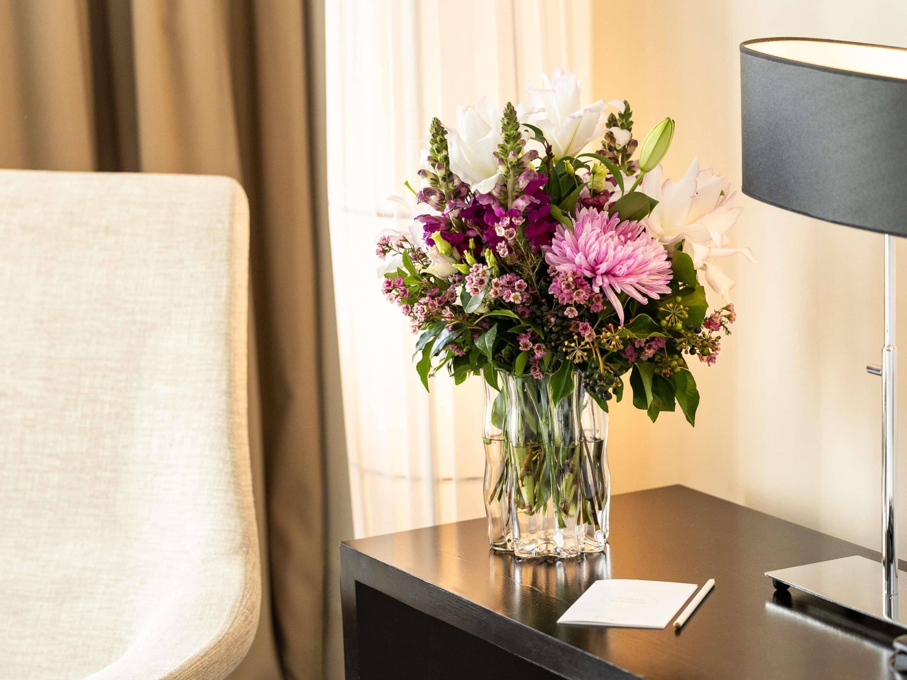 A hotel room with a bed, a nightstand with a vase of flowers, a lamp, and a window with curtains.