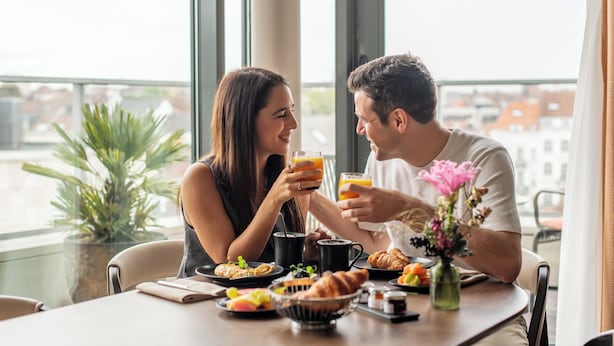 A couple toasting with juice at a table with breakfast items, a plant, and a city view. - Sofitel Brussels Europe (Brussels)