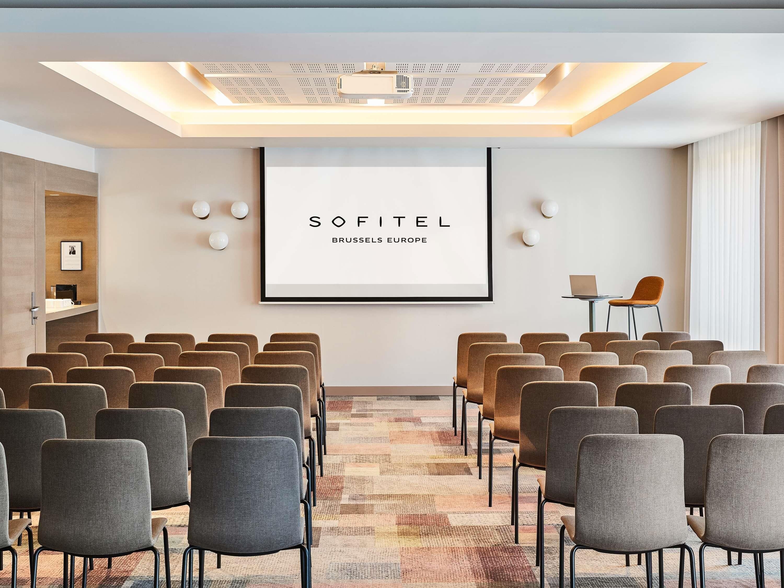 Une salle de conférence avec des rangées de chaises, un écran de projection et un panneau encadré.