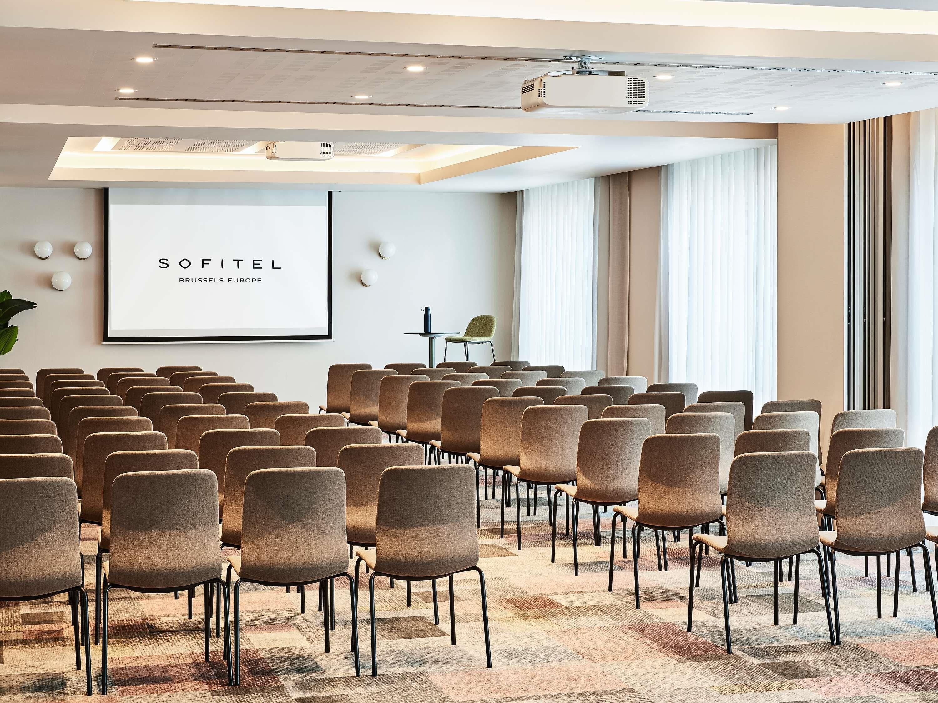 Een vergaderzaal met rijen stoelen, een projecter en een scherm waarop 'SOFITEL BRUSSELS EUROPE' staat.