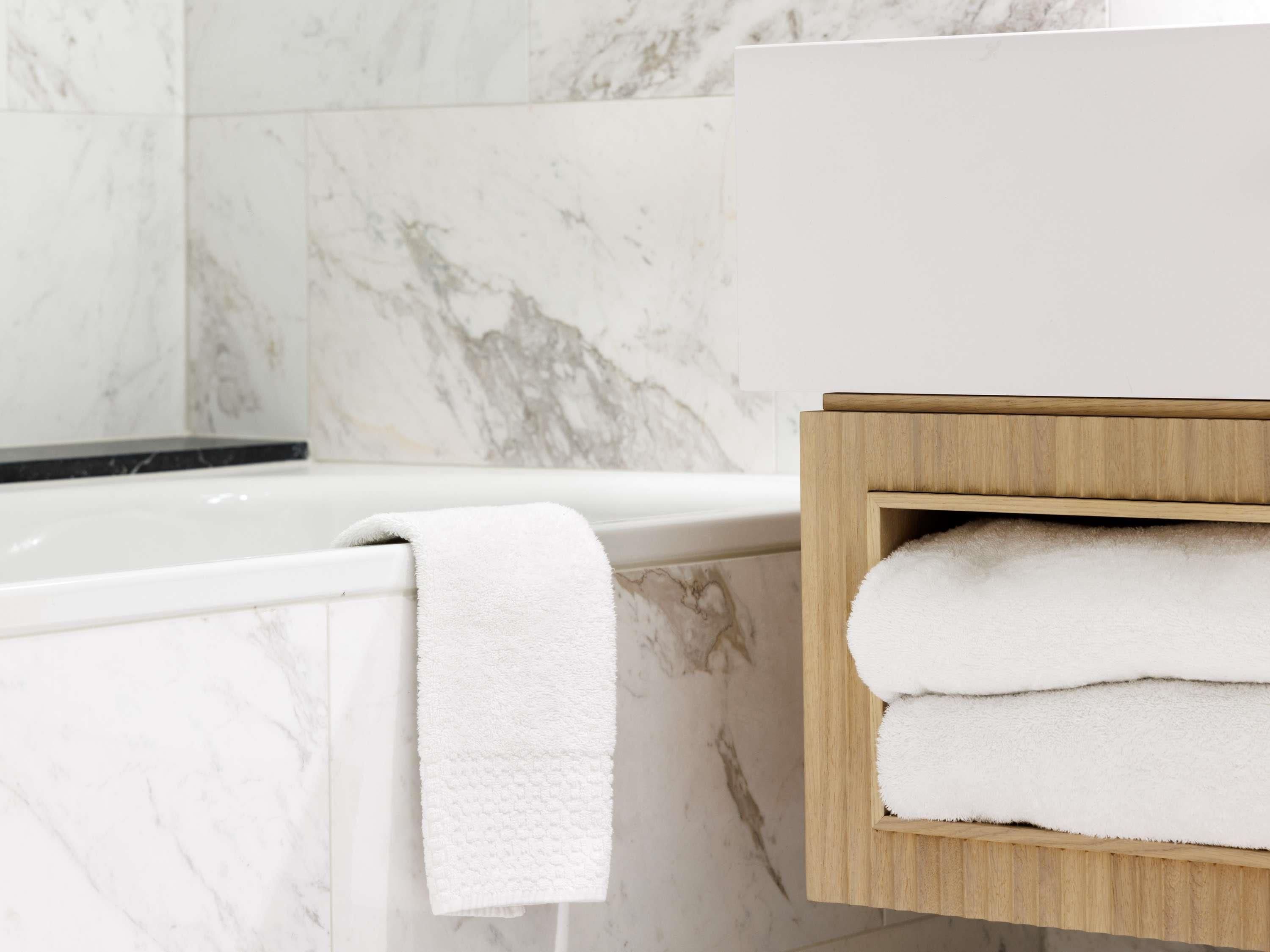 A bathroom with a white marble wall, a white bathtub, and a wooden towel rack with folded towels.