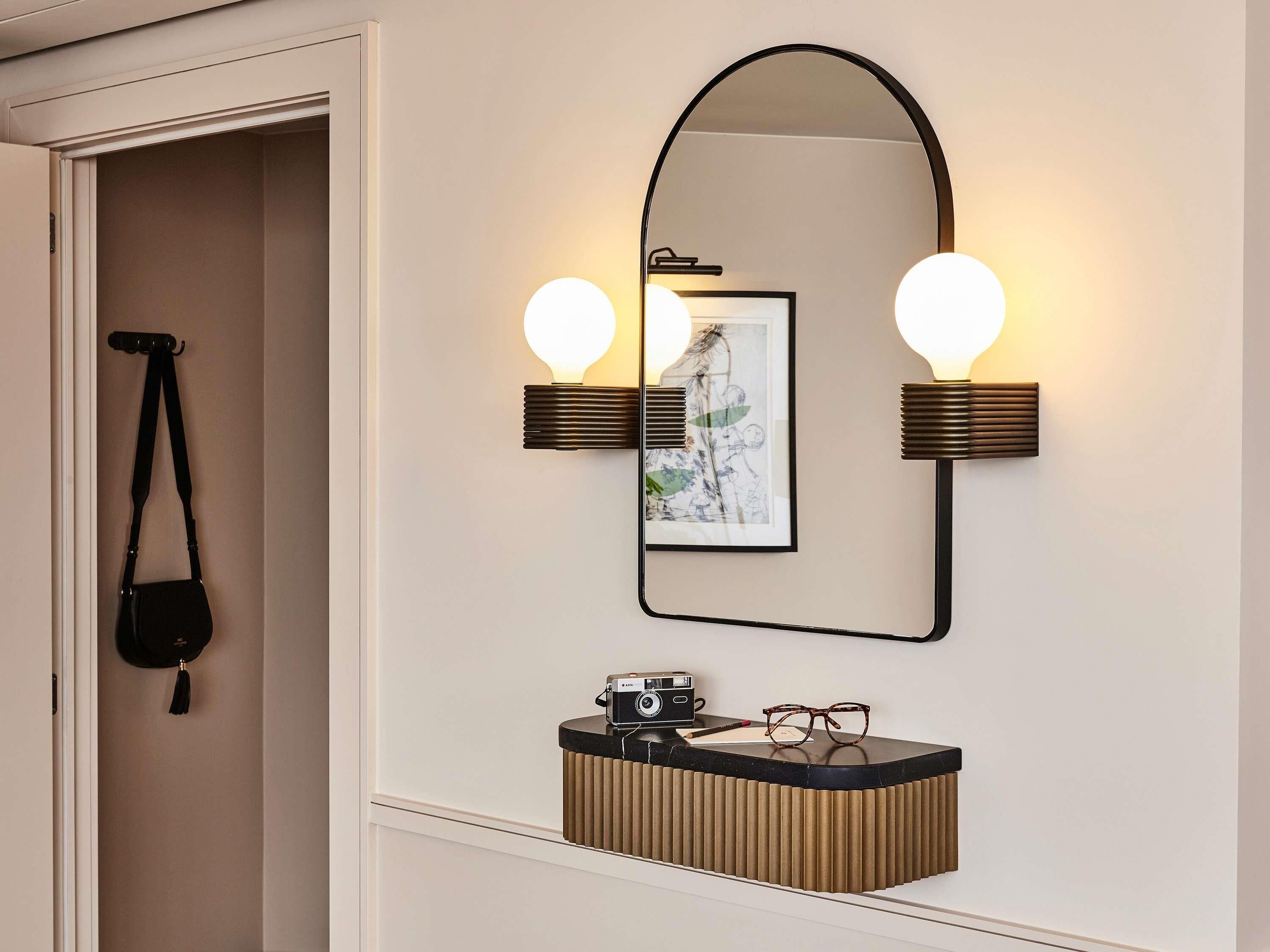A bathroom with a mirror, wall-mounted lights, a framed picture, a vanity with a camera, and a hanging bag.