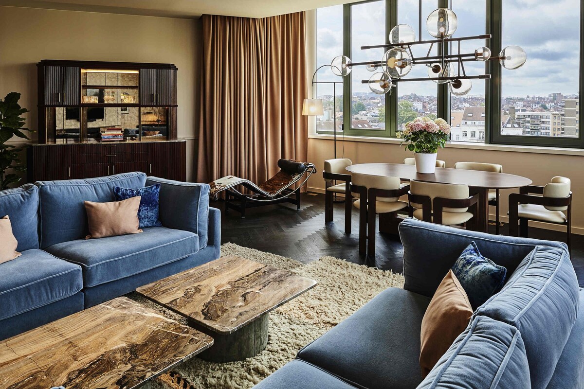 En moderne stue med sofaarrangement, sofabord, spisebord med stole, hængestol og udsigt til byen gennem store vinduer.