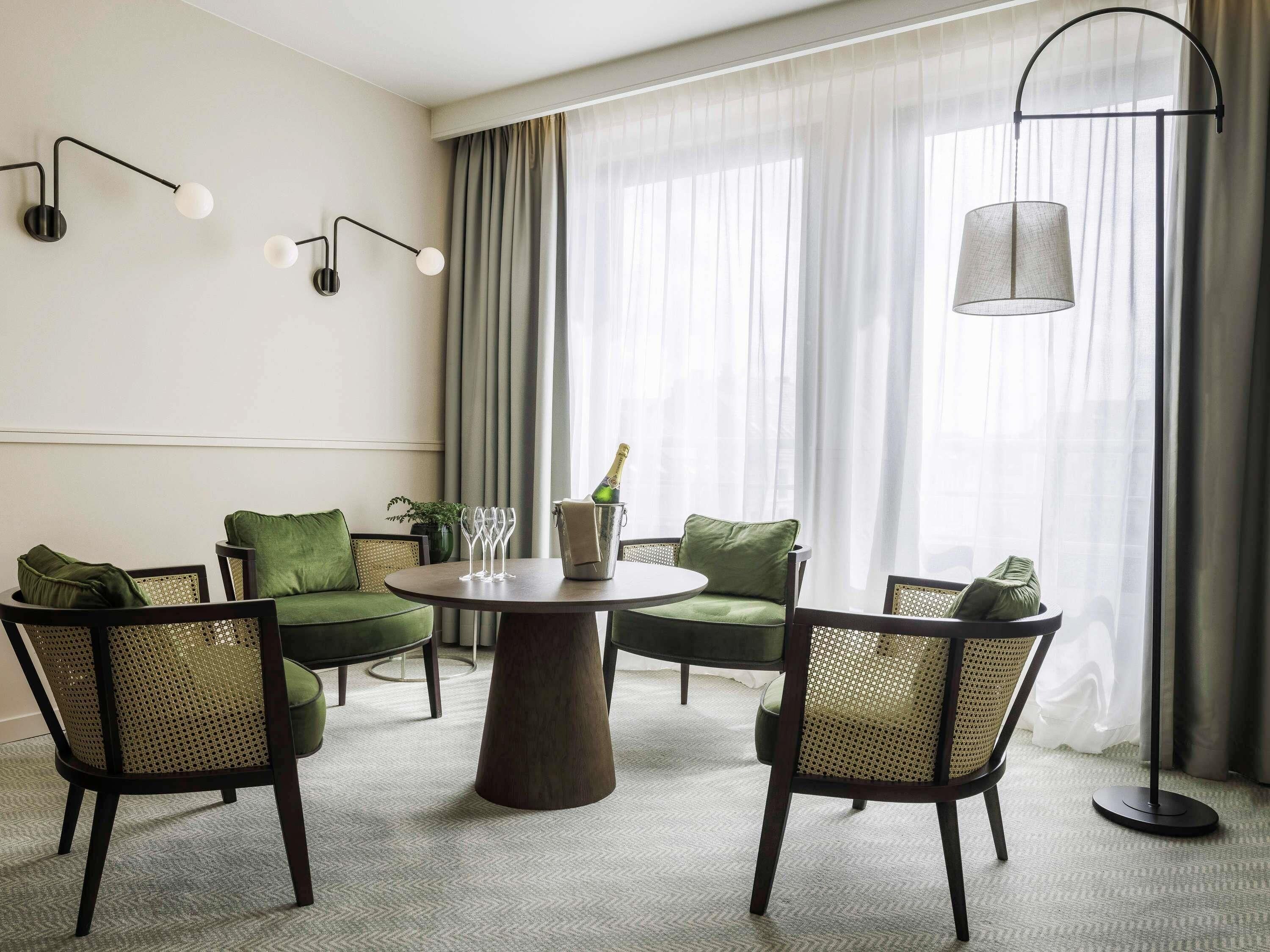 Habitación de hotel con una mesa redonda, cuatro sillas, una lámpara de pie y una ventana con cortinas.