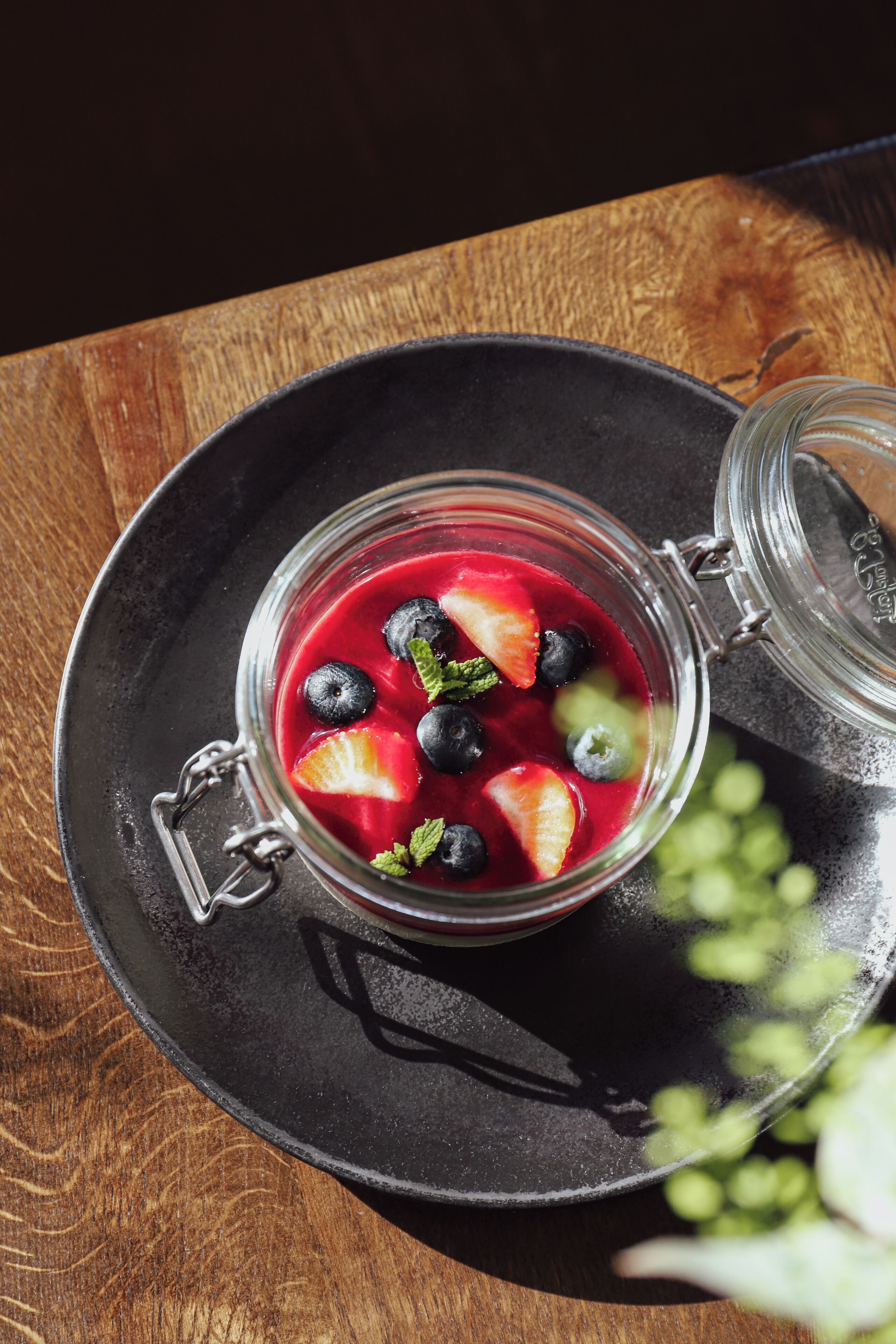 Un frasco de vidrio con tapa metálica que contiene una mezcla de frutos rojos con arándanos y fresas, sobre un plato negro sobre una mesa de madera.