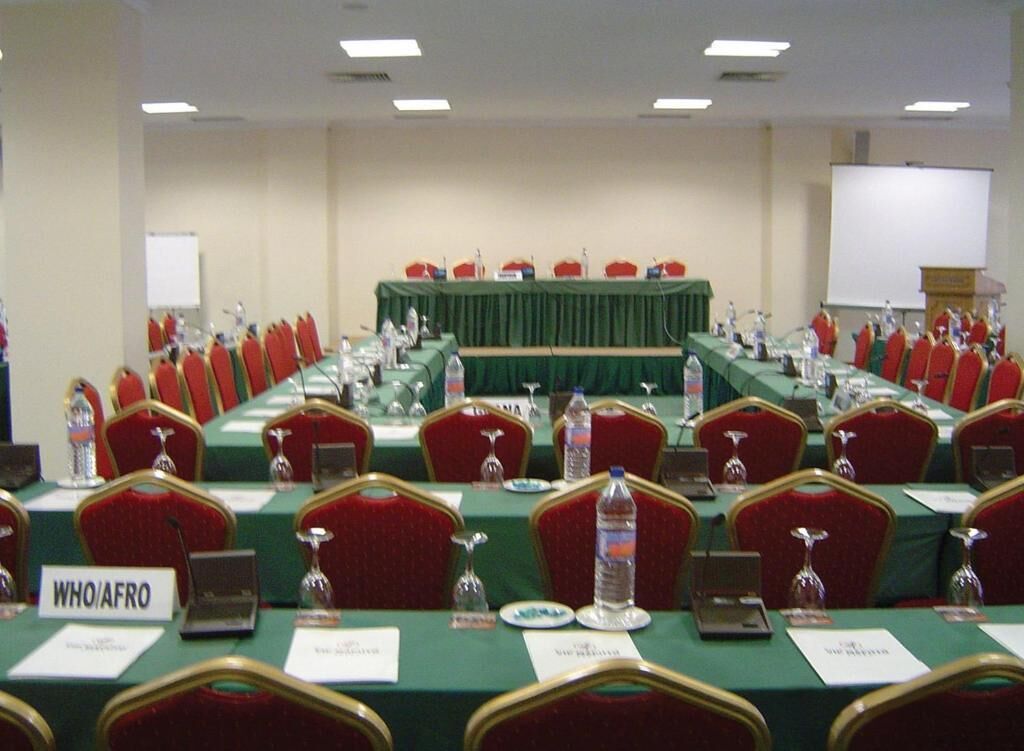 A conference room with rows of red chairs, green tables, nameplates, and a podium.