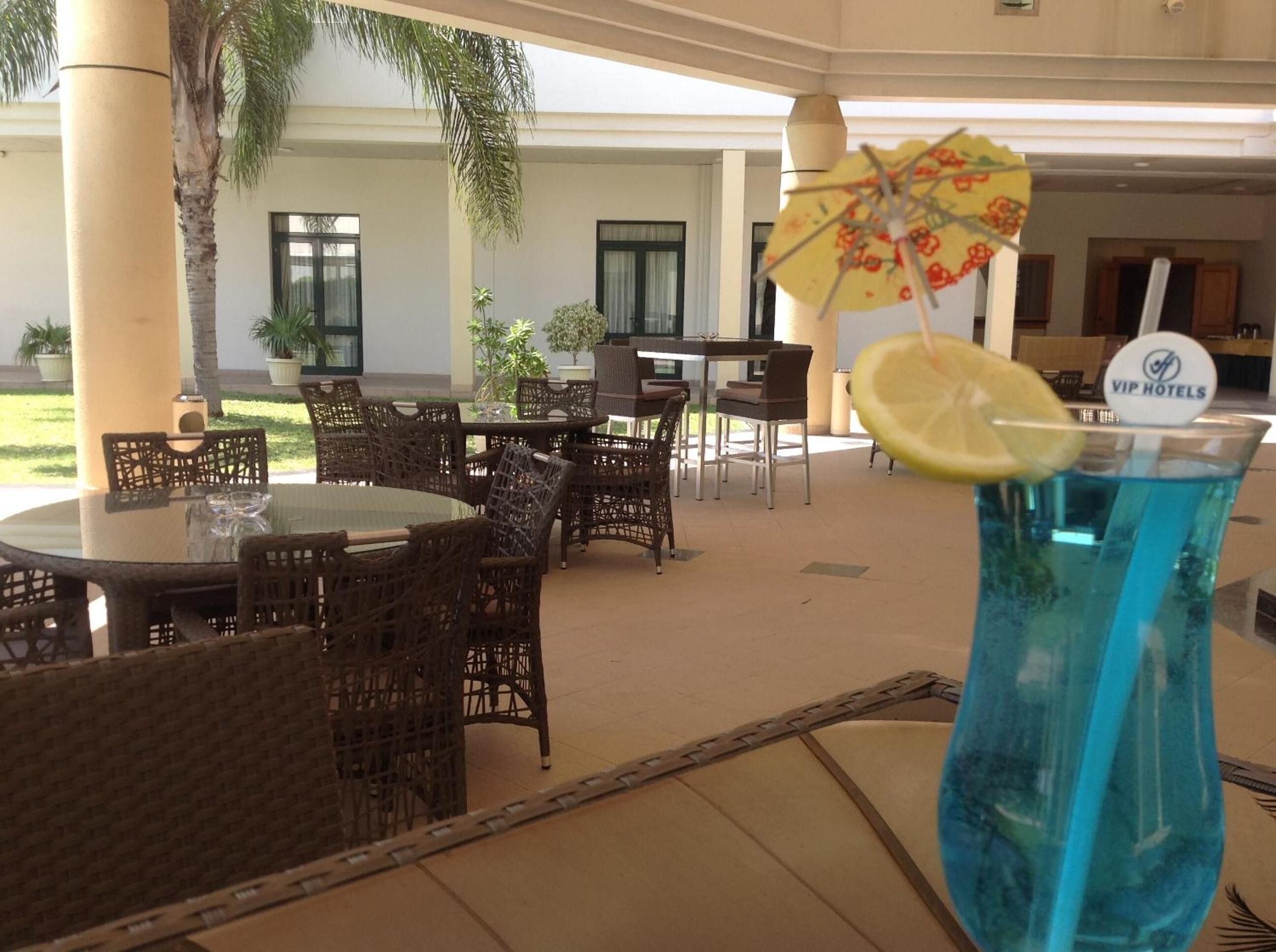 Vestíbulo de hotel con asientos al aire libre, una planta tropical y un vaso de bebida azul con una rodaja de limón.