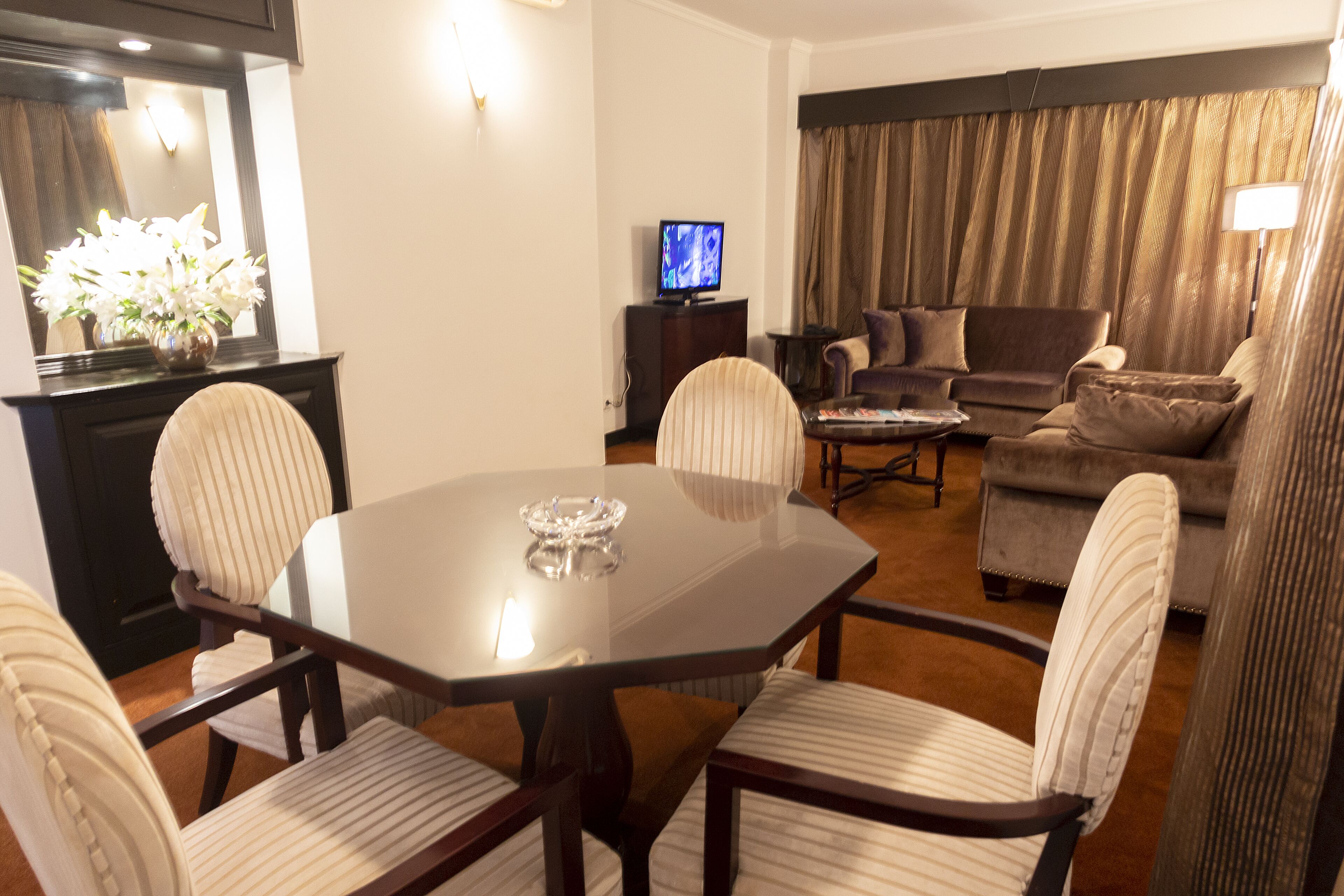 Un área de comedor con una mesa de vidrio, cuatro sillas tapizadas, un televisor sobre un mueble bajo y un sofá al fondo.