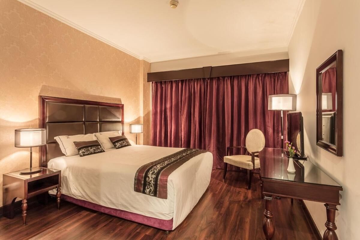 A hotel room with a large bed, a nightstand with a lamp, a desk with a mirror, and a chair.