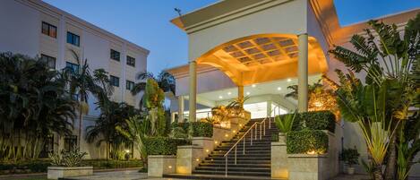 A modern hotel entrance with a covered staircase, palm trees, and a well-lit pathway.