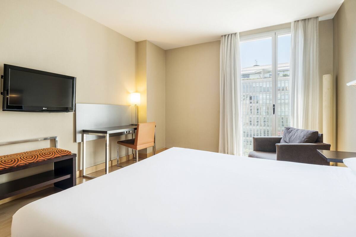 A hotel room with a large bed, a desk with a chair, a TV mounted on the wall, and a window with curtains.