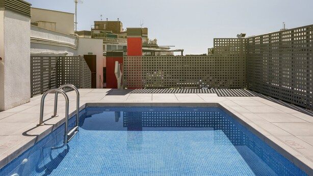A swimming pool with a tiled deck, metal handrails, and a fence. - Hotel ILUNION Auditori (Barcelona)