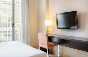 Une chambre d’hôtel comprenant un lit, un bureau avec une chaise, un téléviseur fixé au mur et une fenêtre avec des rideaux.